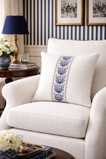 White armchair with a decorative pillow featuring blue patterns, placed in a room with striped wallpaper and framed pictures.